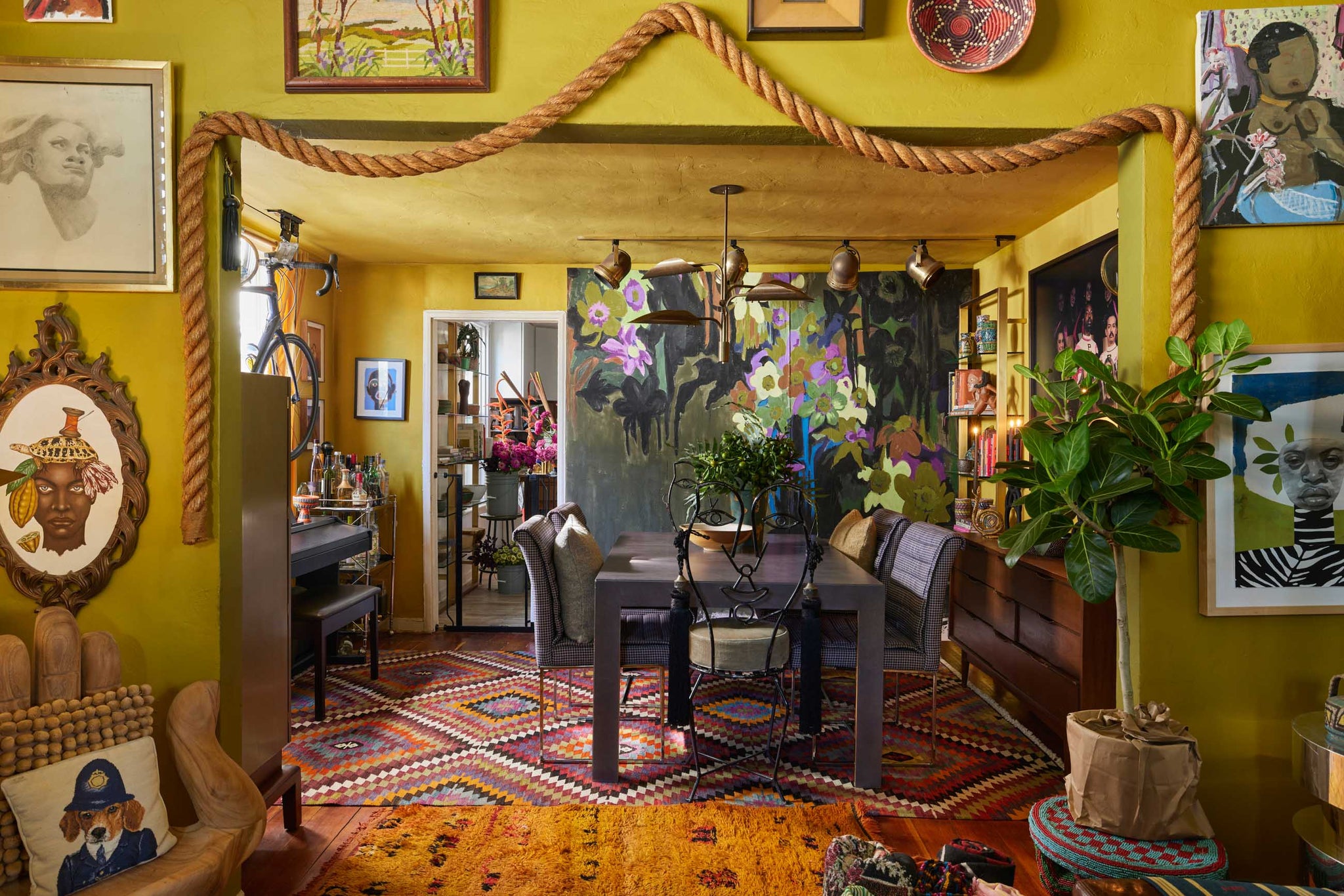 Florist and artist Maurice Harris's dining room has a wooden table with black and white checked chairs on the sides. At the head of the table is an iron chair with a back in the shape of a face. Above the table are bronze spotlights and a bronze chandelier, while a focal floral accent wall is in the back.