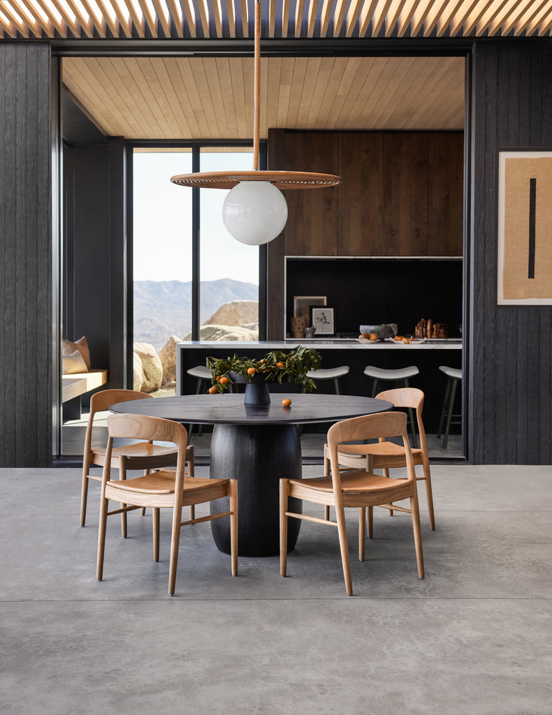 The modern Cassey pendant light has a milky globe attached to a flat rattan disc. The light hangs over a round black wooden dining table with rounded pedestal base and is surrounded by four natural wood curved arm chairs.