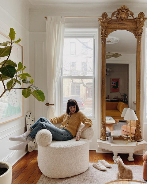 Blogger Mallory Fletchall sits in a round boucle chair. A tall gilded floor mirror stands next to her. An orange cat stares at her.