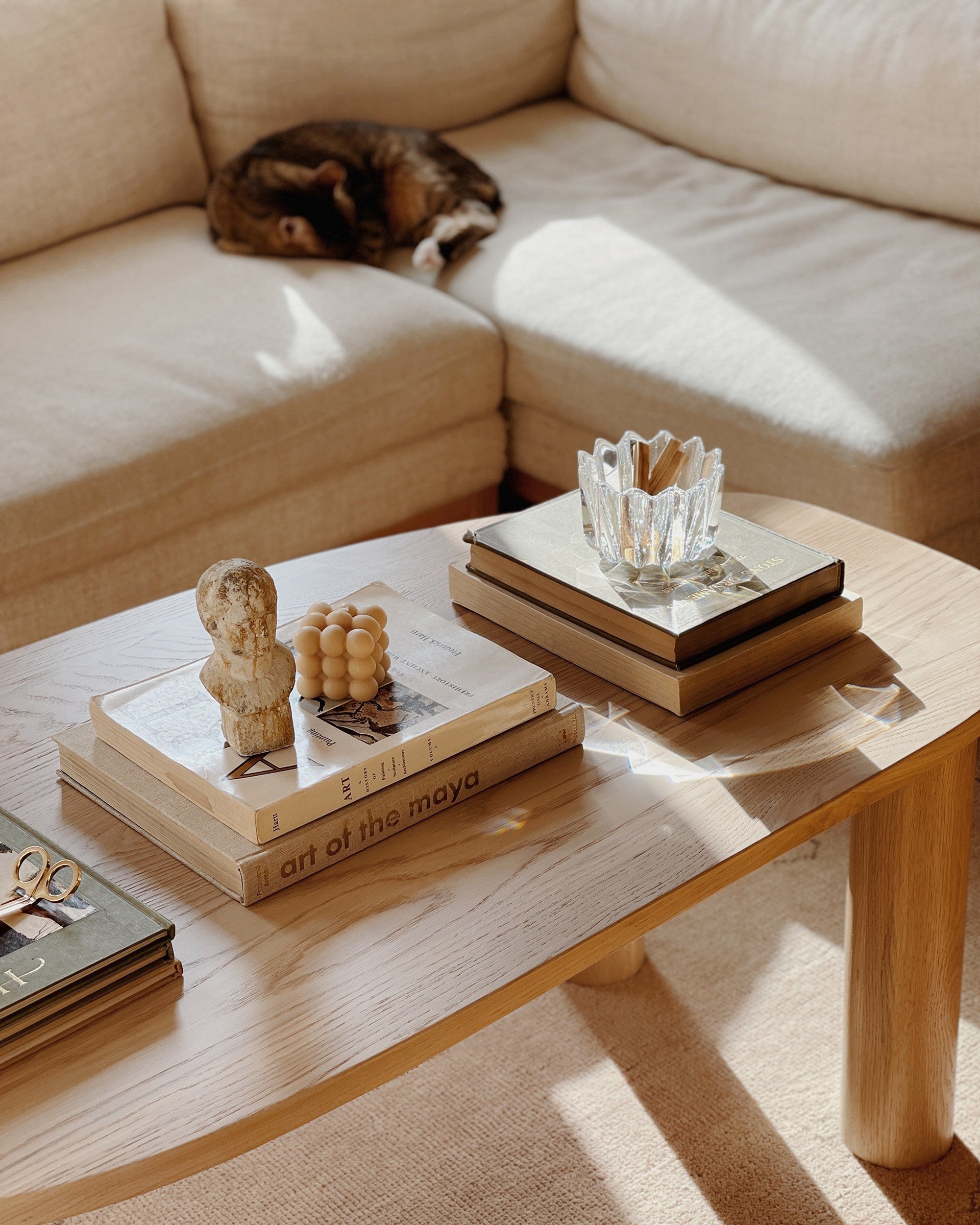 A cat is curled up on an ivory linen sectional sofa. A light wood oval coffee table is in front with stacks of books and decor.