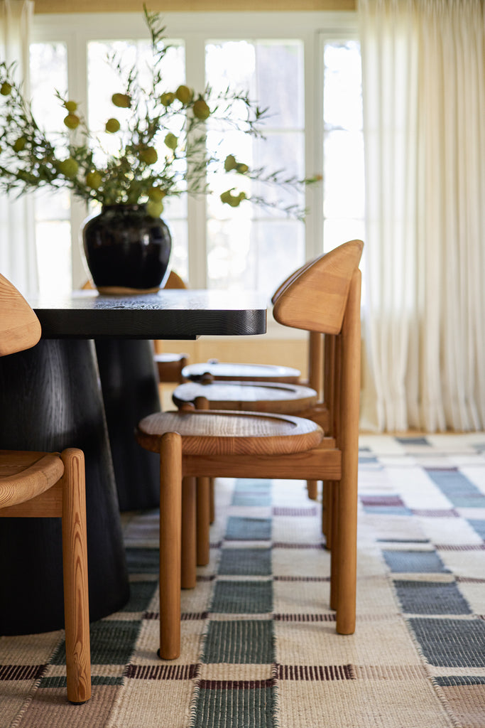 A black modern dining room table us surrounded by light wood curved back dining chairs. The multicolored Marli rug is underfoot.