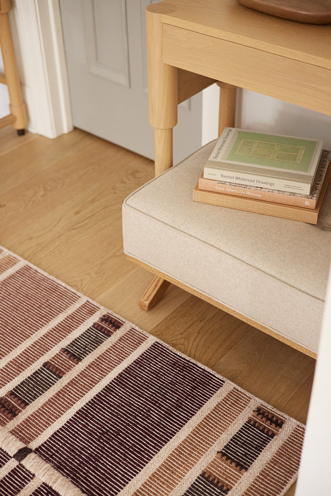 A light wood table with upholstered ottoman sit in front of the multicolored woven Anni rug from Nina Freudenberger's Lulu and Georgia collection.