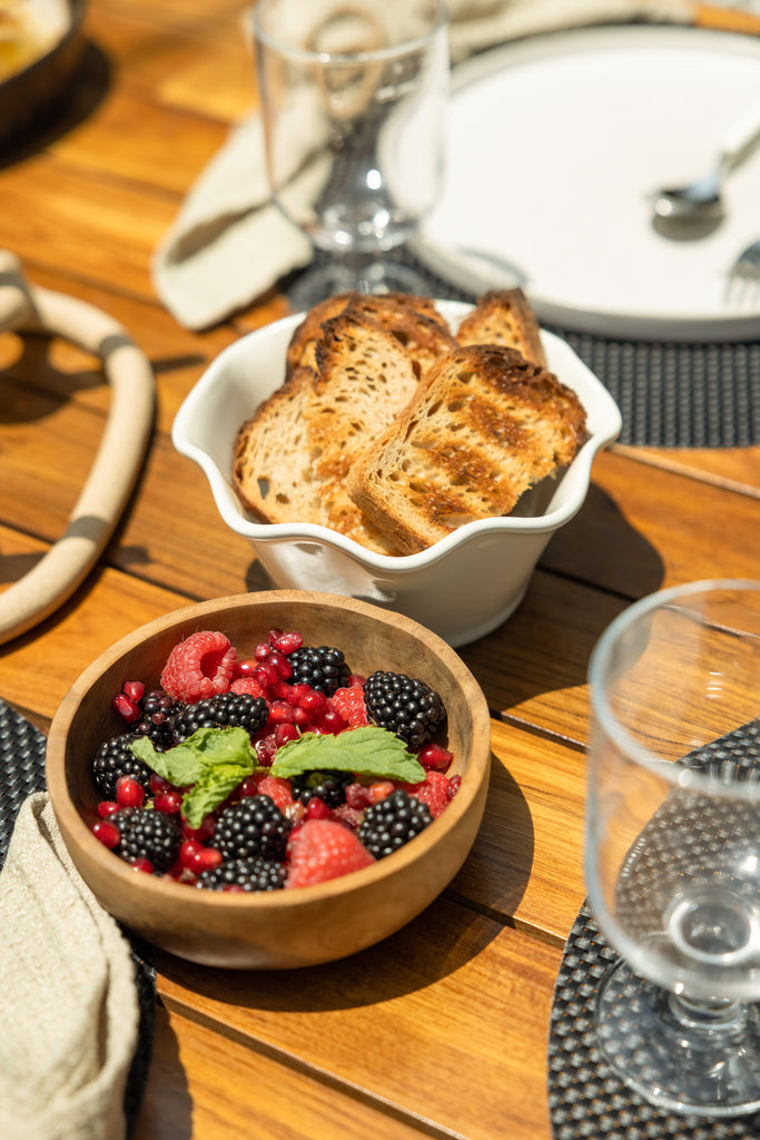 A small wooden bowl of mixed berries sits next to a white ceramic bowl of toasted bread on a teak outdoor table.
