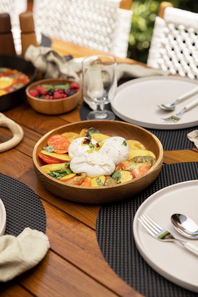 A peach, tomato and burrata salad sits in a wooden bowl between three outdoor place settings with black placemats and white plates.