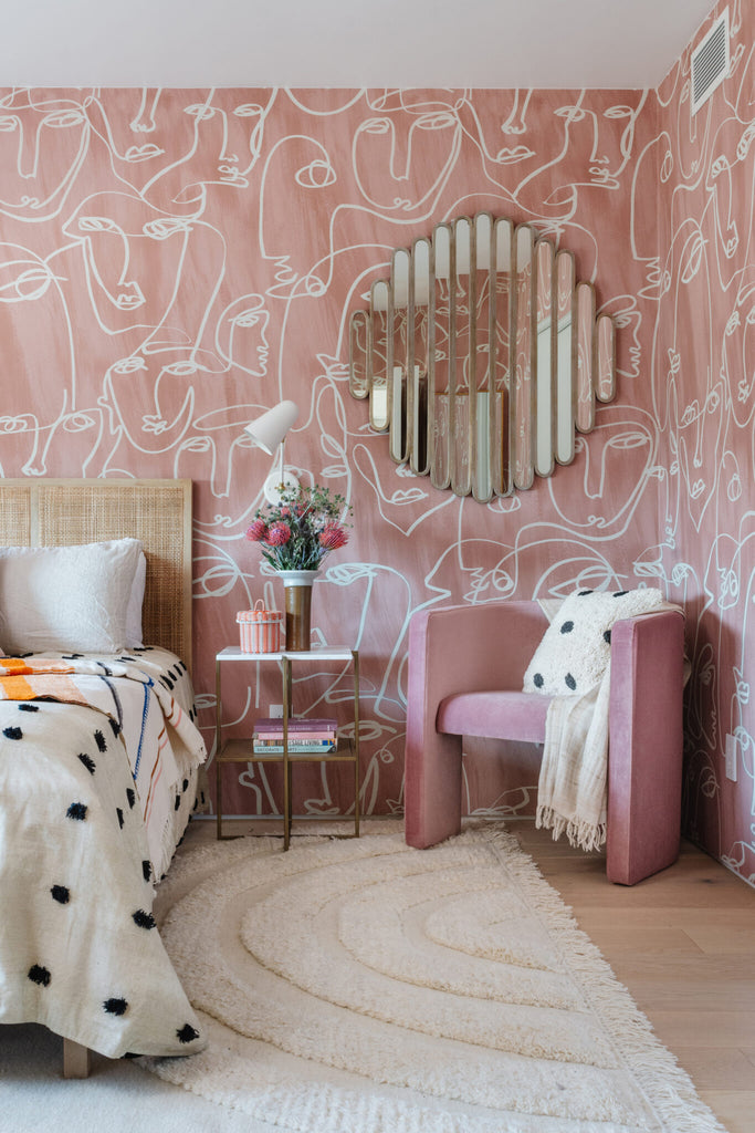 A pink velvet barrel chair sits in the corner of a bedroom with pink wallpaper with abstract faces. Around pieced mirror hangs above the chair, and a light wood and cane Hannah bed with a white duvet with black textured polka dots sits next to it.