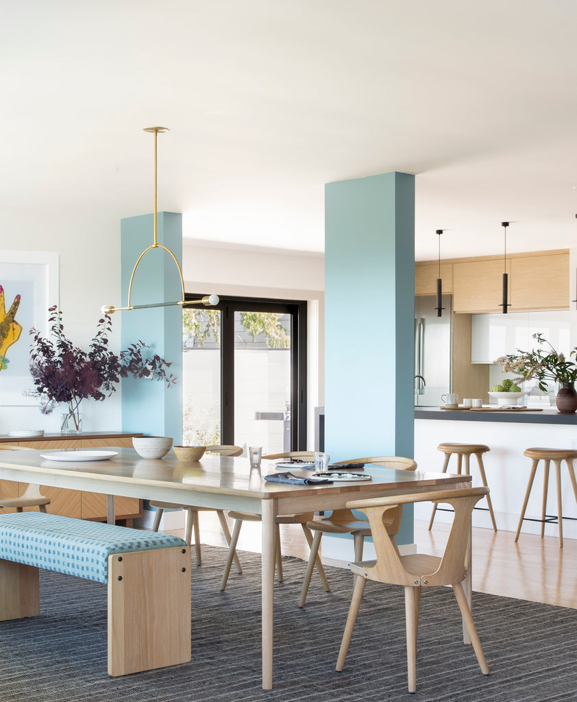 A light wood dining table with light wood dining chairs and a bench upholstered with light blue fabric has a dark gray rug underneath. Behind it is a modern light wood and light blue kitchen.
