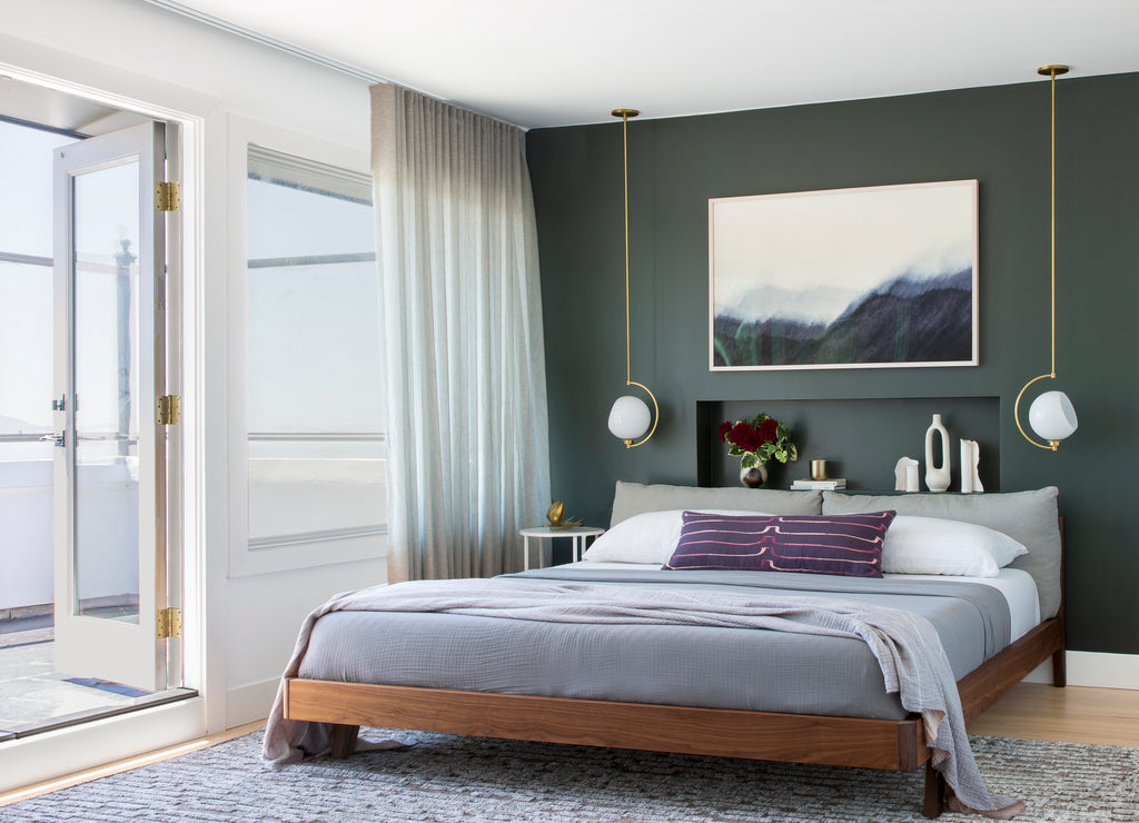 A low wooden platform bed with gray bedding and a purple throw pillow sits against a dark green painted wall with a landscape painting over it. Two gold and frosted glass pendant lights hang on either side of the bed.