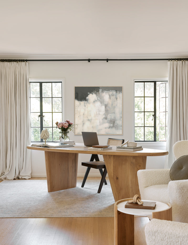 An oval light wood desk  with a laptop, flowers and books sits atop a neutral colored area rug. A black and cane chair sits at the desk, while a black, gray and white abstract painting is on the wall behind it. A boucle accent chair sits near the desk.