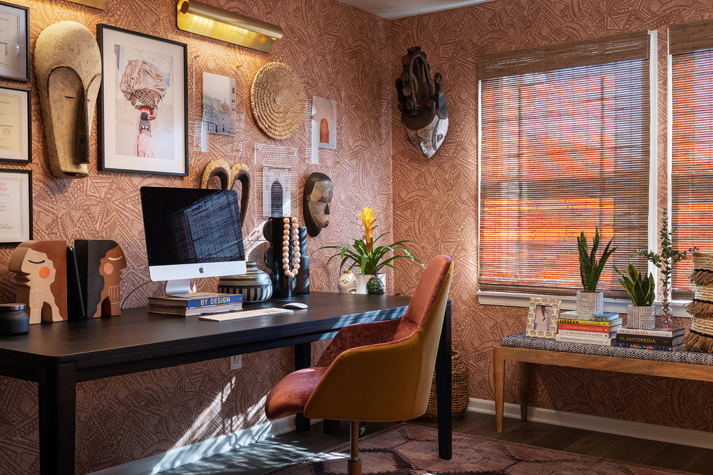 Designer Beth Diana Smith's office has reddish brown Alaari geometric wallpaper, African art hanging on the wall above a dark wood desk with an upholstered chair. A bench with stacks of book and plants sits in front of a window.