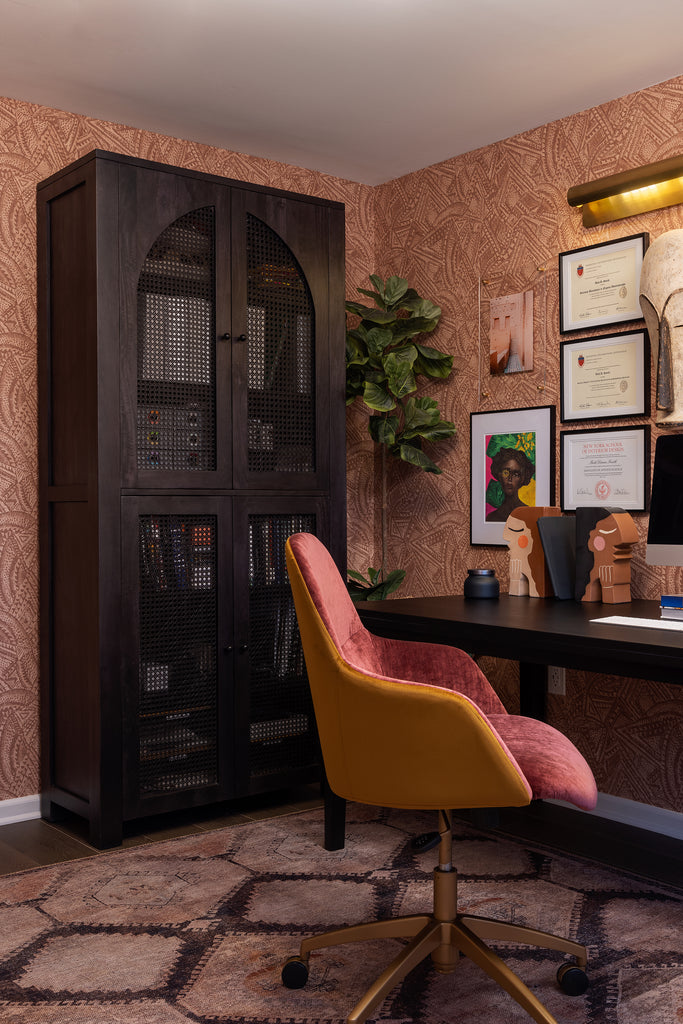 A red and gold upholstered desk chair sits against a dark wood desk. The dark arched Islay storage cabinet stands in the corner, and the reddish brown Alaari geometric wallpaper hangs in the room.