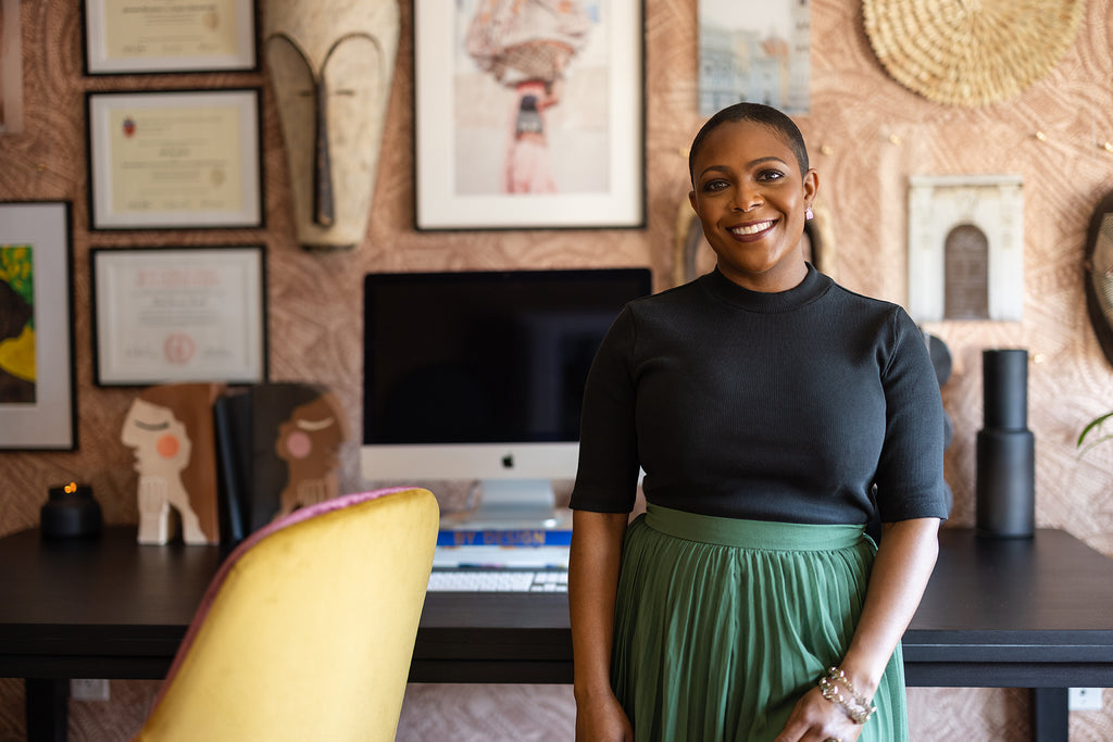 Interior designer and CEO Beth Diana Smith stands in front of her desk in a black shirt and emerald green skirt. Next to her is a gold and red upholstered desk chair. Behind her is a black desk with an Apple computer and hanging art on a red patterned wallpaper.