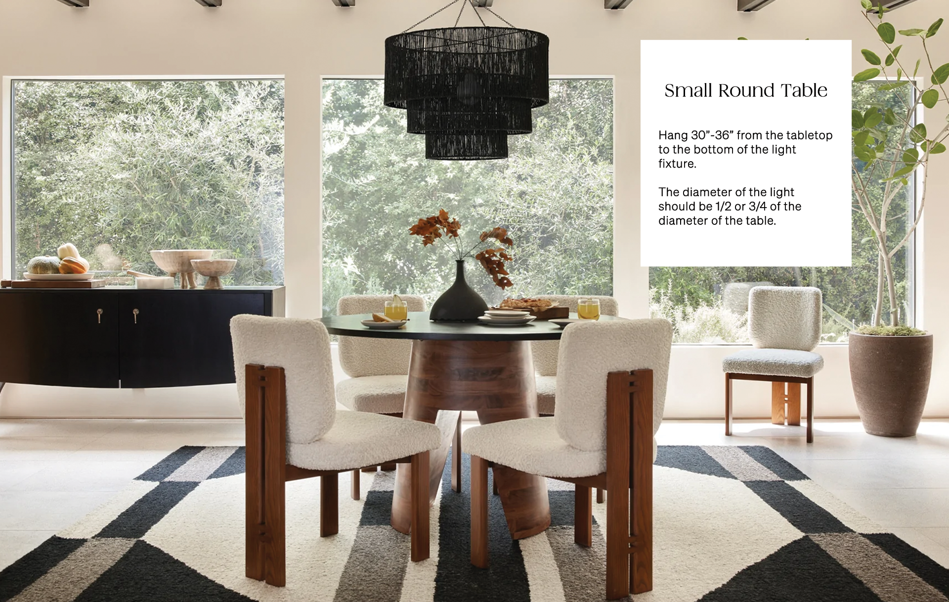 The large black Chavette rattan chandelier hangs over a round pedestal dining table with wood base and black top that is surrounded by four boucle upholstered chairs with wooden frame.