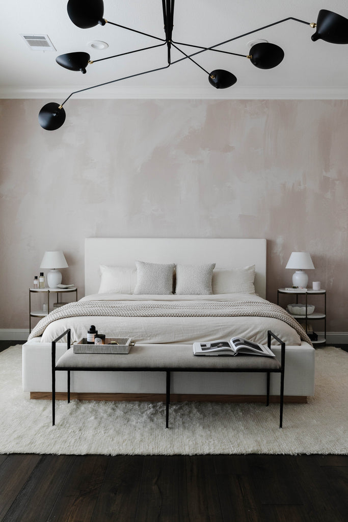 Chriselle Lim's bedroom has a white upholstered bedframe, white bedding, two matching iron and marble rounded nightstands, an iron bench and a modern linear chandelier.