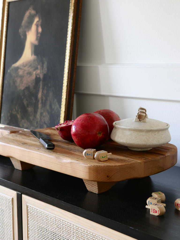 A wooden cutting board with pomegranates and dreidels sits on a black and cane sideboard cabinet.