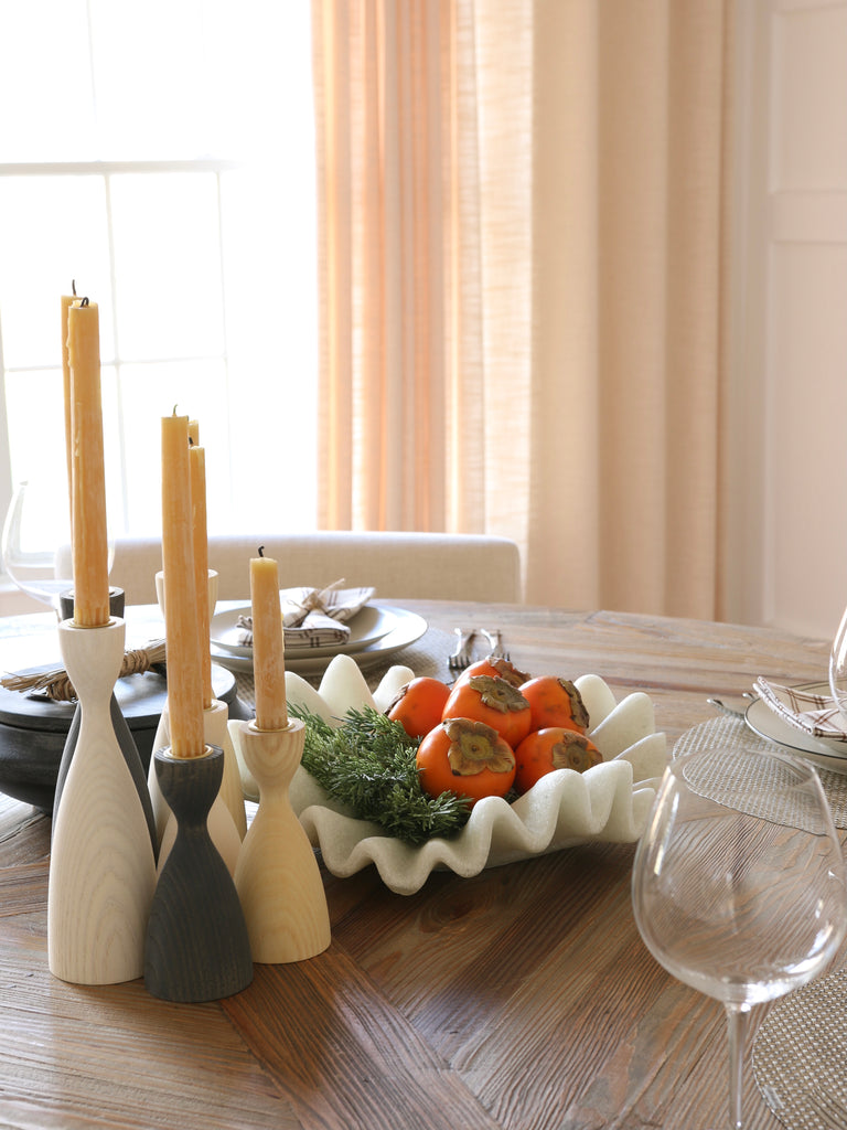 Farmhouse Pottery wooden candlestick holders in different colors and heights sit next to an Arteriors Flora wavy centerpiece bowl holding persimmons on a round wooden dining table.