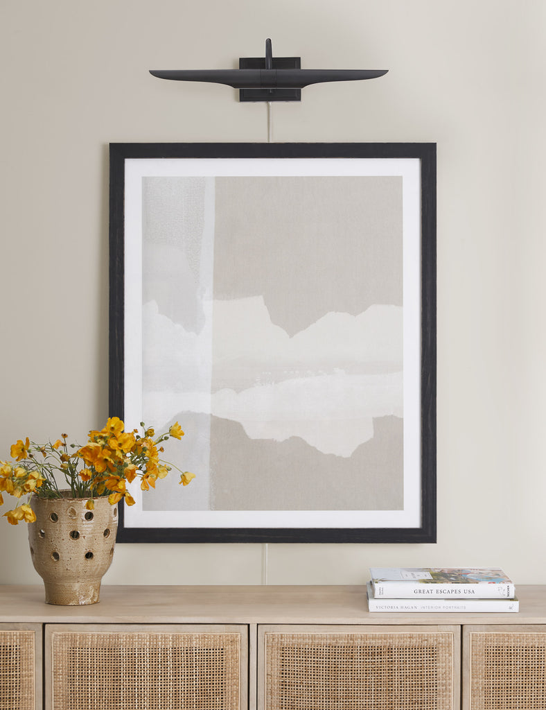 The Regina Andrew Redford Picture Light is a black flat wall light that hangs above an abstract neutral-toned landscape painting, which is hung above a light wood and cane sideboard.