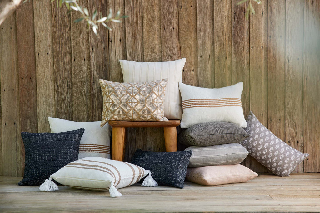 A large stack of throw pillows lays on a wooden floor and against a wood paneled wall. The pillows are in hues of cream, gray, blush, navy and mustard.