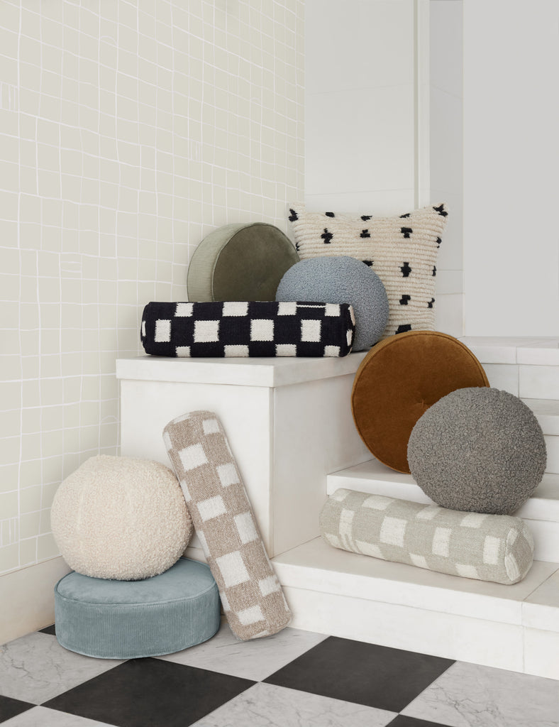 The Irregular Checkerboard Bolster pillow and Boucle Ball Pillow sit on a white pedestal in various colors. The pillows were designed by Sarah Sherman Samuel for Lulu and Georgia.