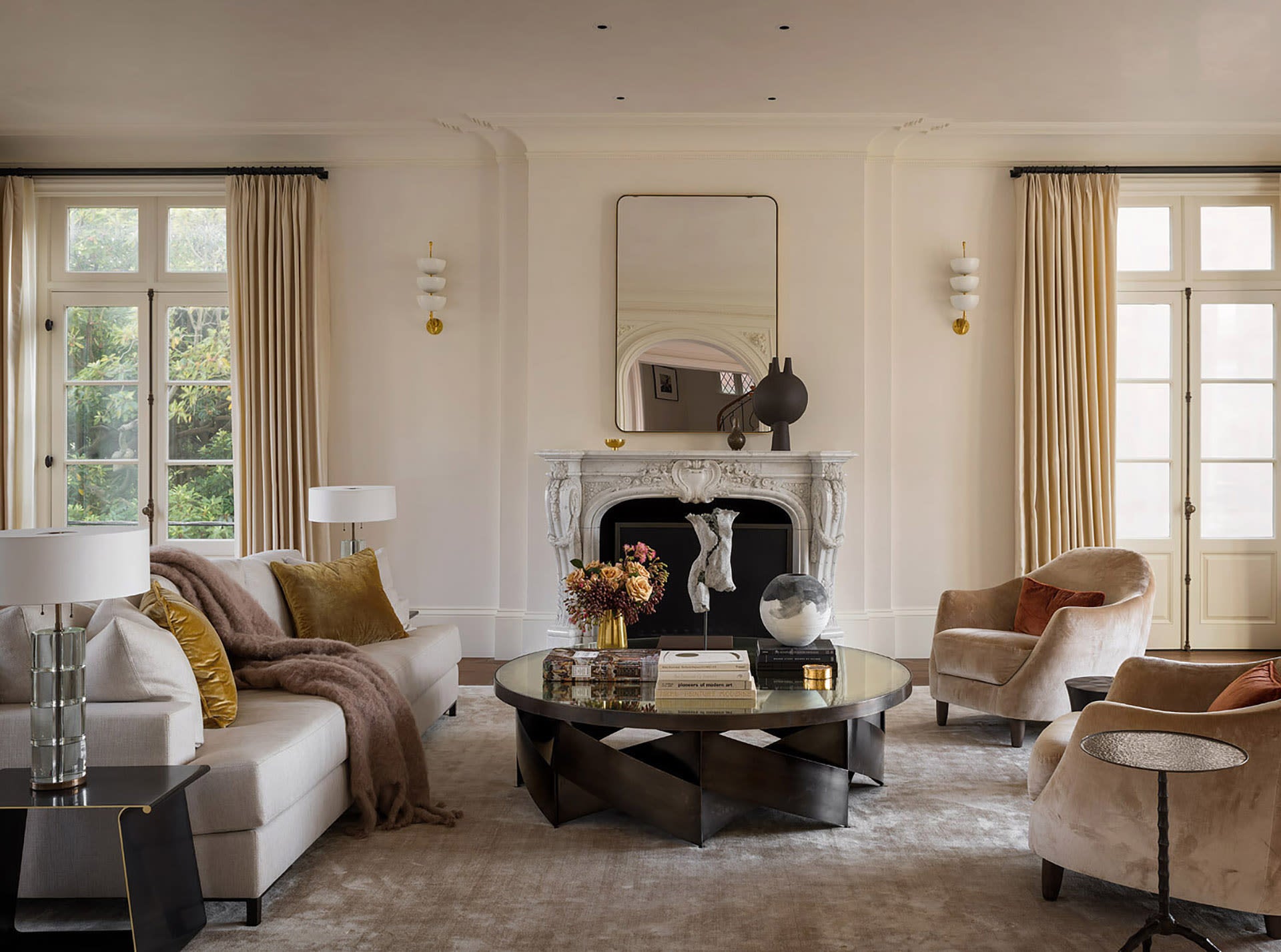 A classic living room has an ornately carved stone fireplace 
 with a modern rectangular mirror above, an ivory sofa with gold throw pillows, two taupe velvet rounded accent chairs, and a large round black coffee table with mirrored top.