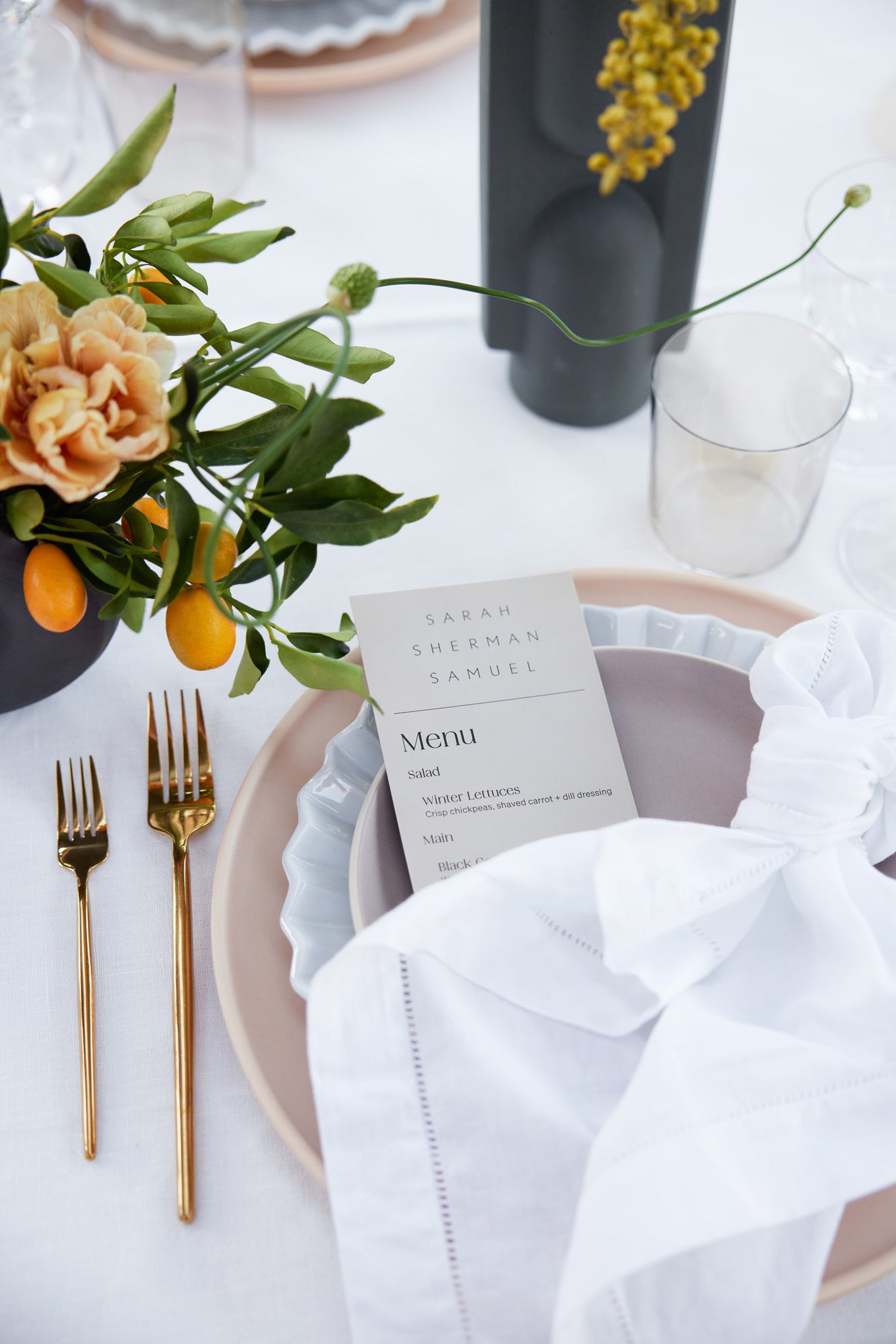 A place setting with layered plats, gold flatware, a white knotted napkin and a menu are part of a celebratory dinner for the Sarah Sherman Samuel x Lulu and Georgia collection. A menu is tucked under a napkin.