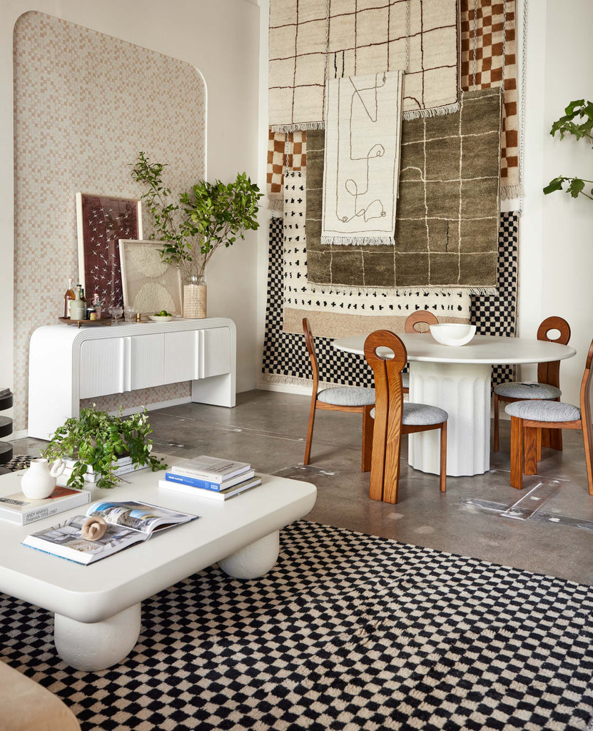 Area rugs from Sarah Sherman Samuel's Lulu and Georgia collection hang on the wall of a pop-up shop in LA. In front of the wall is the white Merritt Sideboard with a minibar, paintings and vase holding branches on it, and a round white pedestal dining table surrounded by five curved wooden dining chairs. In front is a black and white checked area rug with a white curved square coffee table with books and magazines.