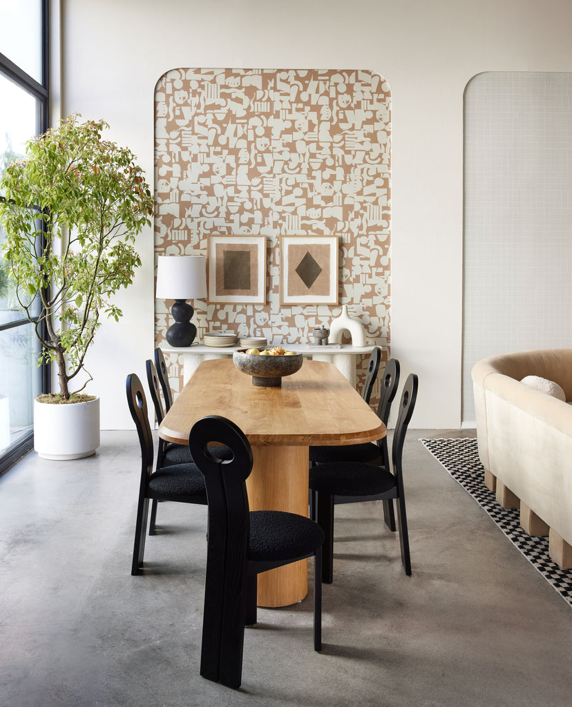 The Organic Shapes Wallpaper by Sarah Sherman Samuel hangs behind a dining room set-up at a pop-up shop in LA. Two geometric paintings hang on the wall and an oval wooden dining table surrounded by seven black curved dining chairs sit in front of the wall.