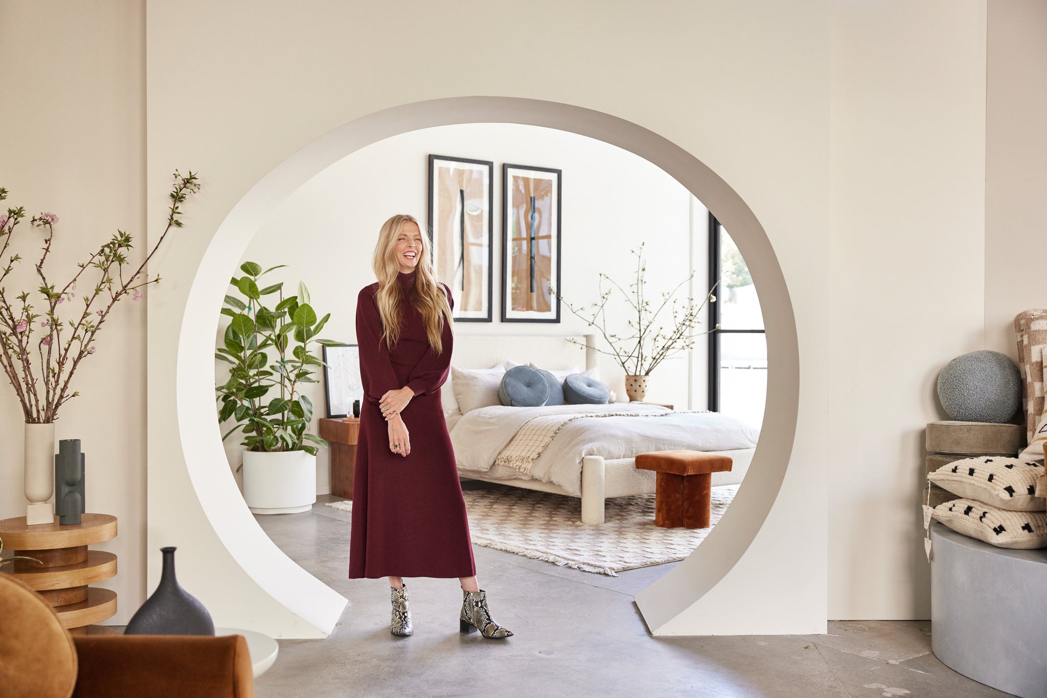 Interior designer Sarah Sherman Samuel stands in a rounded bedroom entryway in an LA pop-up shop. Behind her is the white boucle Hyvaa bed with neutral linens and flanked with plants. Stacks of pillows and vases line the entryway.