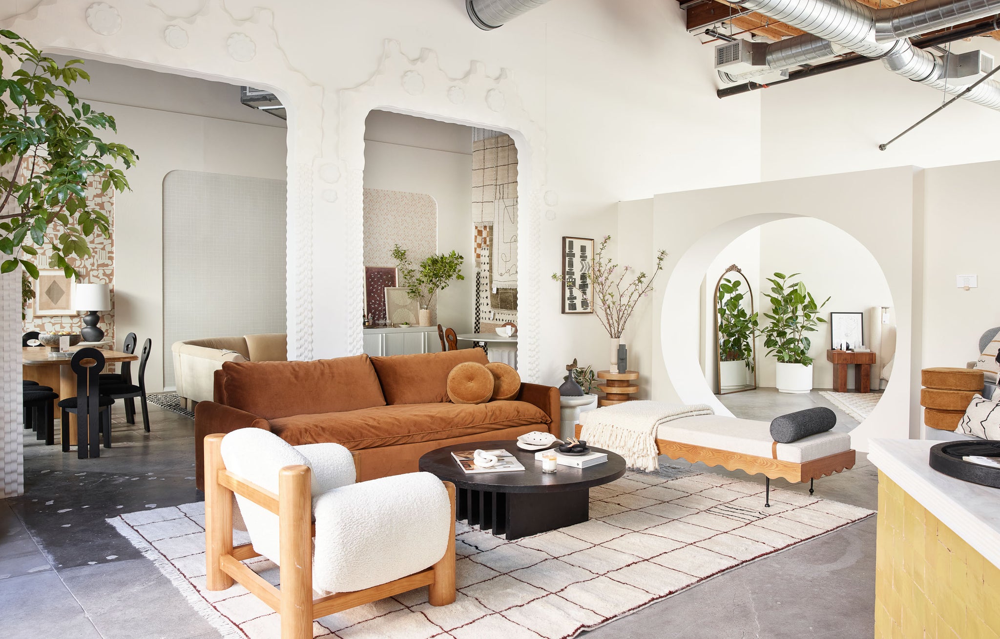 A modern home set-up is displayed in the Sarah Sherman Samuel x Lulu and Georgia pop-up in LA. A brown velvet sofa is flanked by a modern rippled daybed and a sherpa-covered accent chair. Behind the living room is a dining room with wooden dining table and black curved chairs and a seating area with a light taupe sectional sofa.