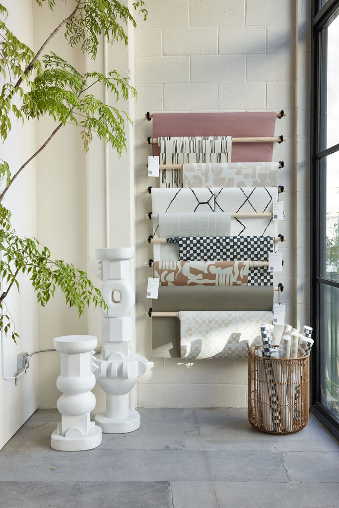 Nine of Sarah Sherman Samuel's modern wallpapers are draped on hanging rods in a pop-up shop in LA. In front is a rattan basket holding rolls of wallpaper and next to it are the Toivo pedestals.