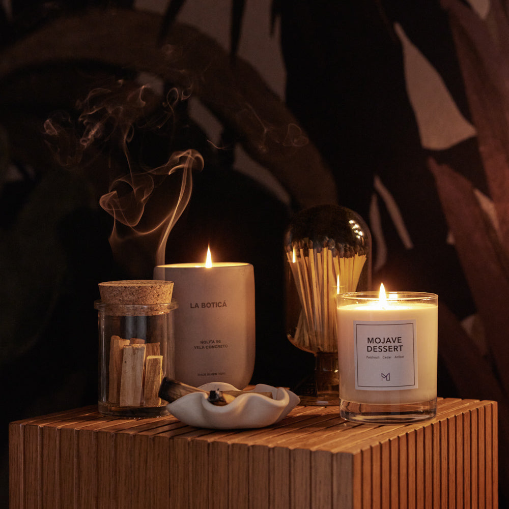 A lit La Botica candle and Mojave Dessert candle sit atop a wooden cube with a match cloche and class canister of Palo Santo wood.