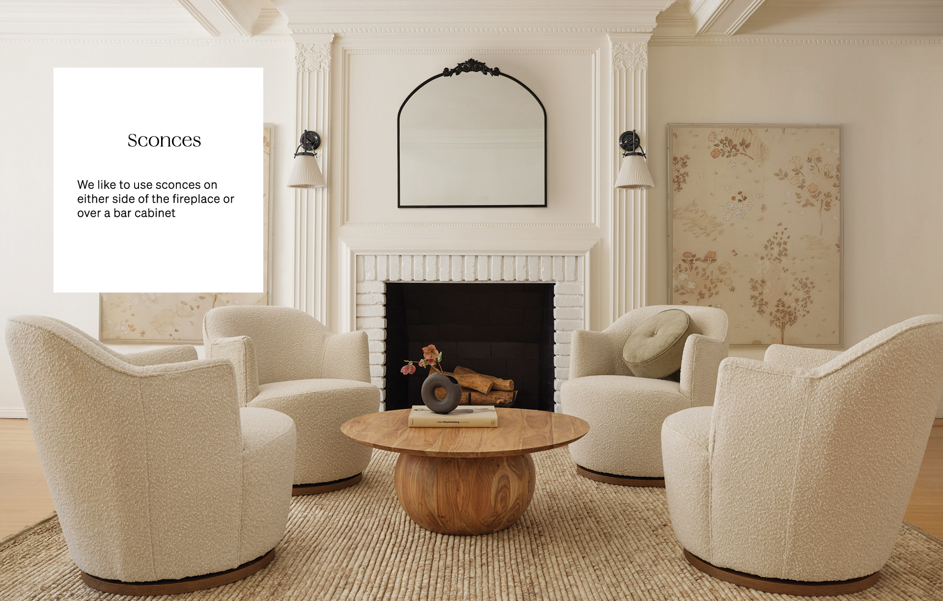 Two wall sconces hang on either side of a fireplace with a black framed arched mirror above the fireplace. Four rounded ivory barrel boucle chairs surround a round wood pedestal coffee table.