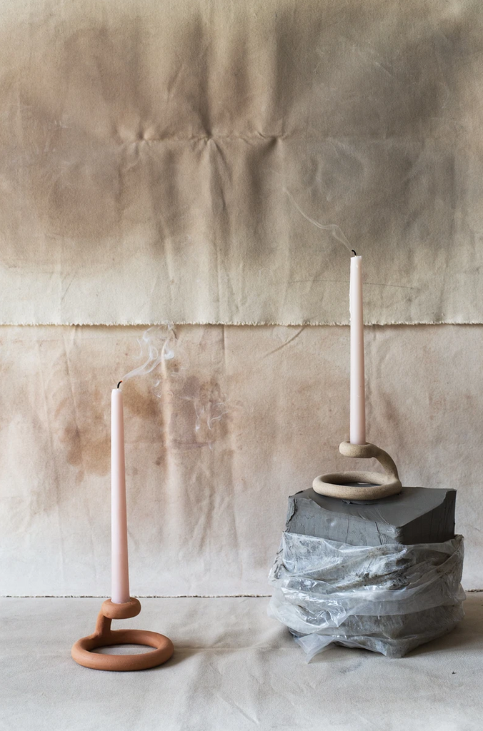 Two coiled taper candle holders holding recently extinguished tapered candles sit against a rough neutral-colored fabric background.