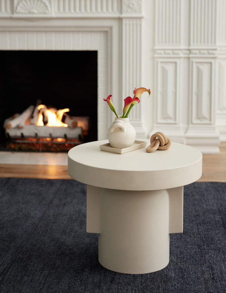 A round white sculptural side table holds a white ceramic vase with three lilies and a decorative knot object.