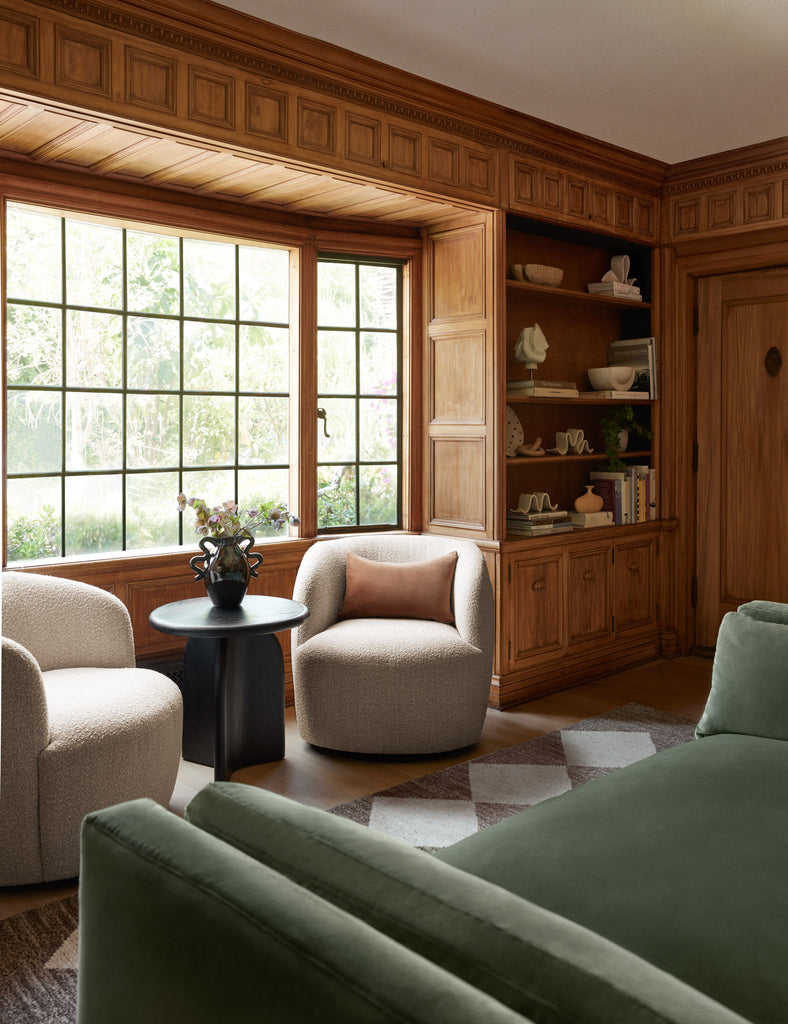 Two neutral boucle barrel swivel chairs sit across from a green velvet sofa in a wood-paneled library. A small round black accent table sits between the chairs and a brown and white diamond patterned area rug flanks the area.