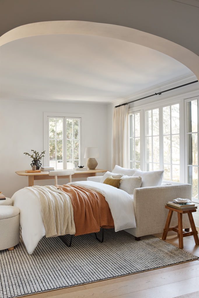 A pulled out sleeper sofa bed with neutral bedding sits in front of a wooden desk and on top of a cream and black checked rug.