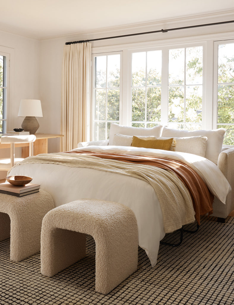 A pulled-out sleeper sofa bed has neutral bedding, sits in front of a window, and has two ivory boucle stools at the foot of the bed.