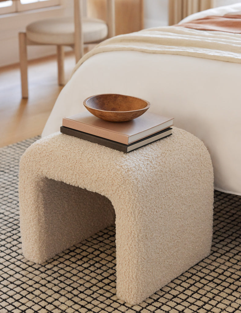 A curved ivory boucle stool sits atop a cream and black check rug and has a two books stacked with a wooden bowl on top.