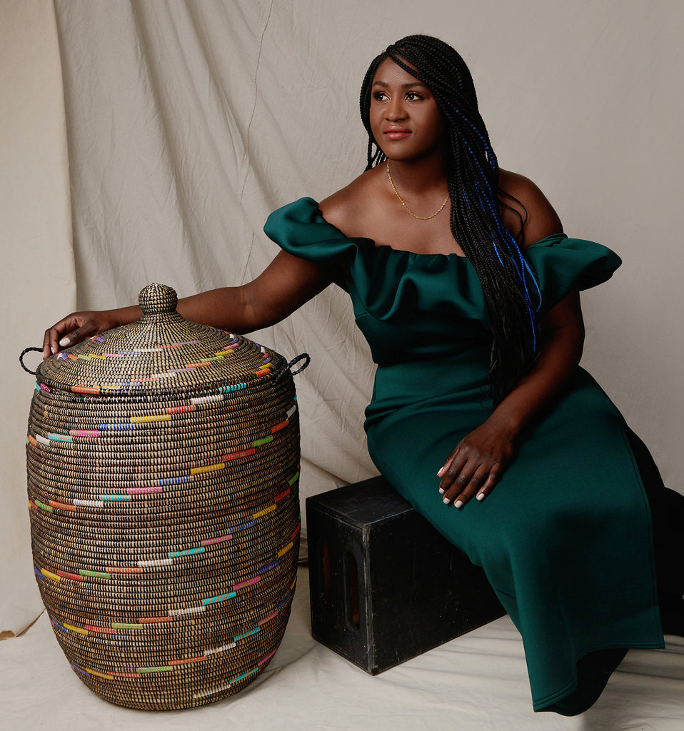 Expedition Subsahara founder and fiber artist Sofi Seck wears a dark green emerald dress and leans against one of her large brown and multi-colored baskets with a lid.