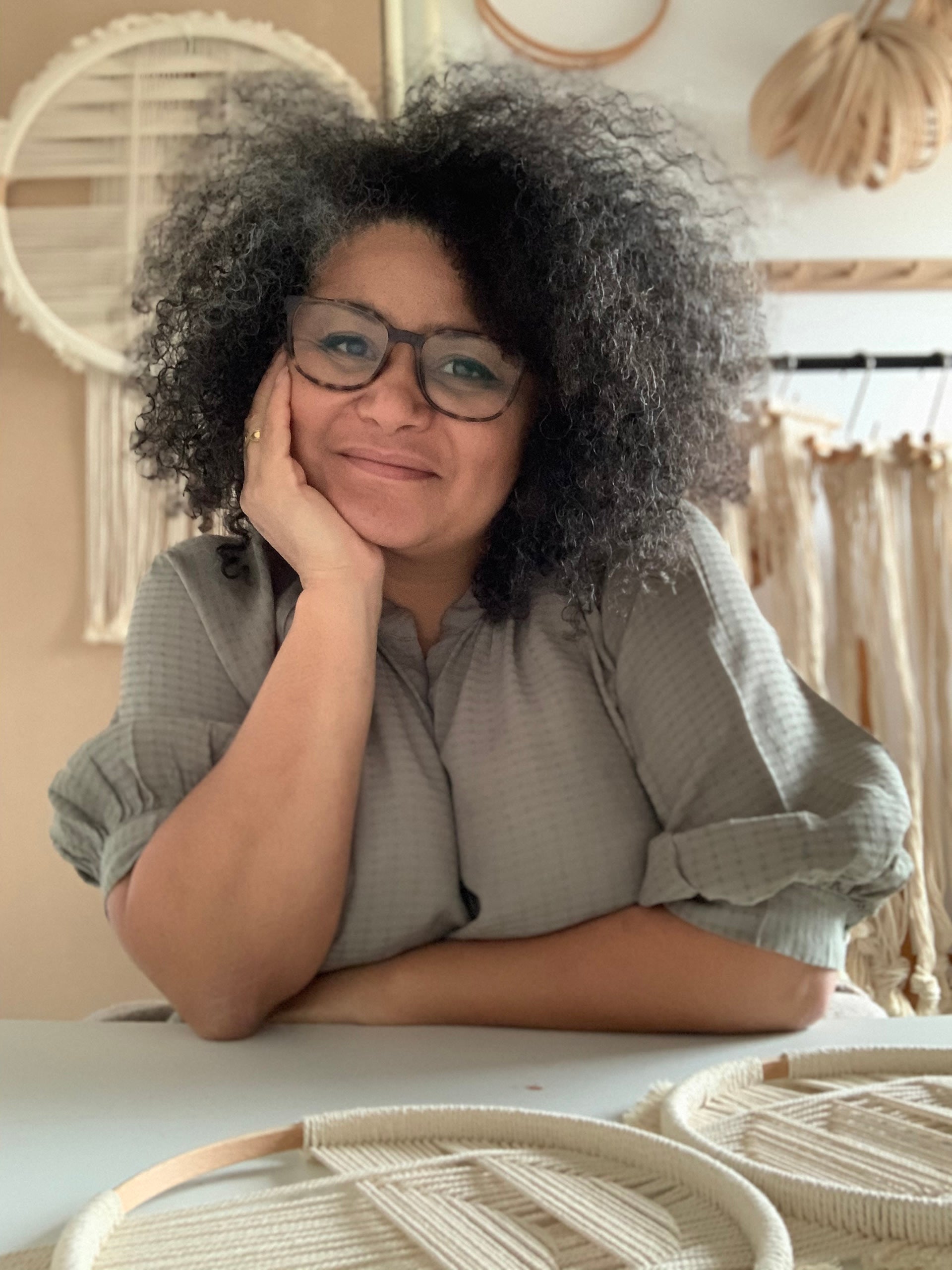 The founder of Studio Nom, Nompumelelo, leans on a counter with her head in her hand. On the counter and behind her are some of her woven wall hangings.