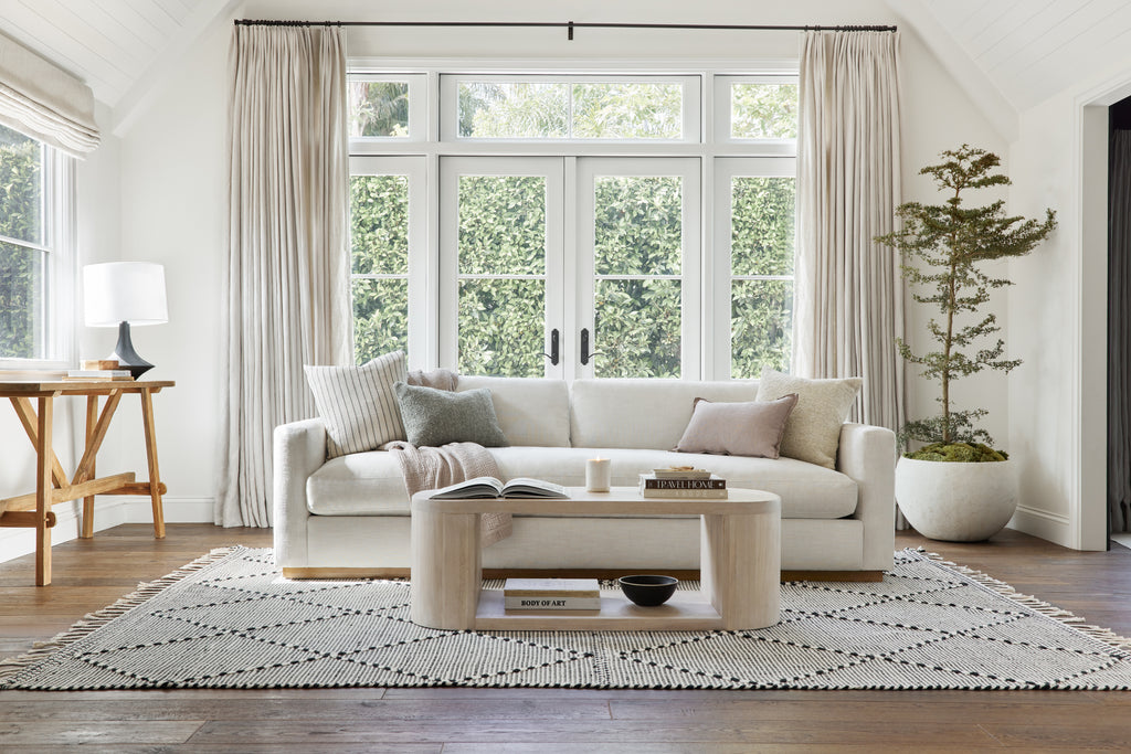 A white linen sofa sits atop the Taza woven rug in black and white. A rounded light wood coffee table sits in front of the sofa.