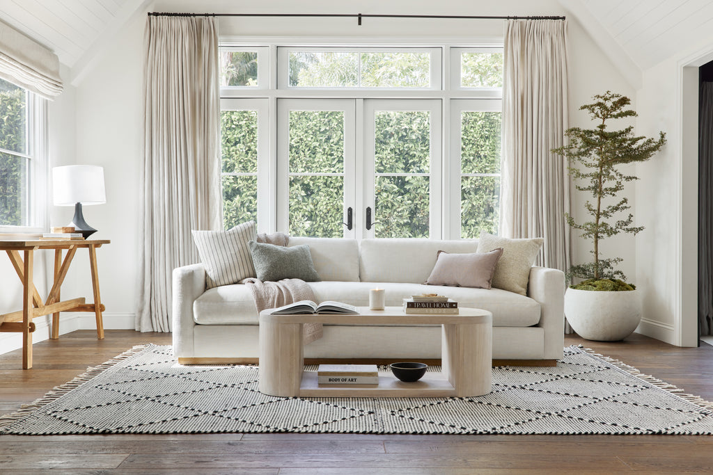 The black and cream striped square Vdara throw pillow, a gray boucle lumbar pillow, the rose colored Arlo linen lumbar pillow and an ivory boucle pillow sit on a squared arm ivory sofa in a soft traditional living room look.