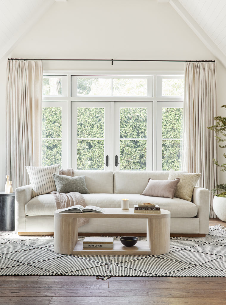 The Walden ivory linen performance fabric sofa sits atop the durable black and white woven Taza area rug. In front of the sofa is a rounded light wood coffee table.