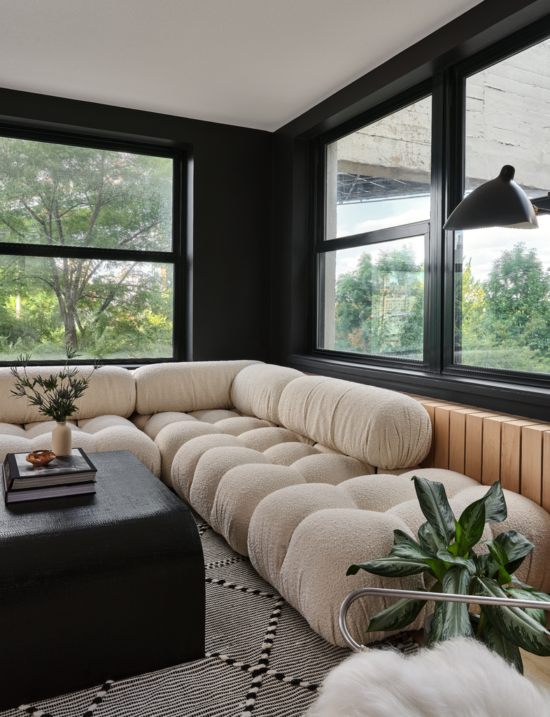 A low oatmeal boucle tufted sectional sofa sits in the corner in front of windows. A square black wooden coffee table sits in front holding a stack of books.