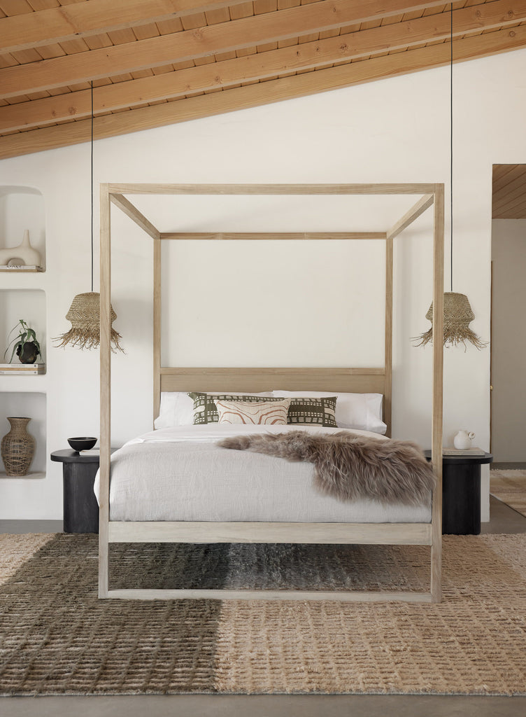 A light wood four poster canopy bed has neutral bed linens, dark olive throw pillows and a fuzzy blanket. On either side of the bed hangs a woven pendant light above a small dark wood night stand. A woven area rug in neutral hues is below the bed.