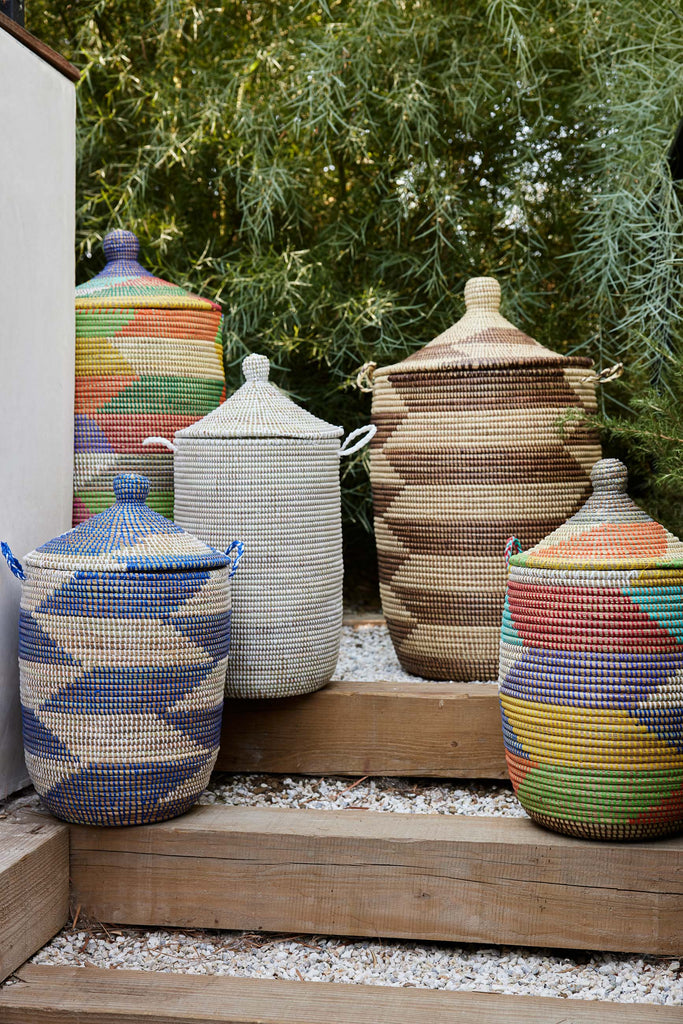 Five Expedition Subsahara storage baskets with lids in a variety of colors sit outside on wood and gravel steps.