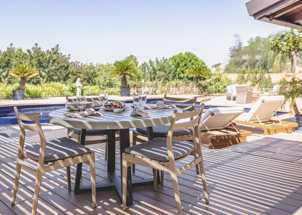 A round concrete dining table surrounded by light wood dining chairs sit outside by a pool.