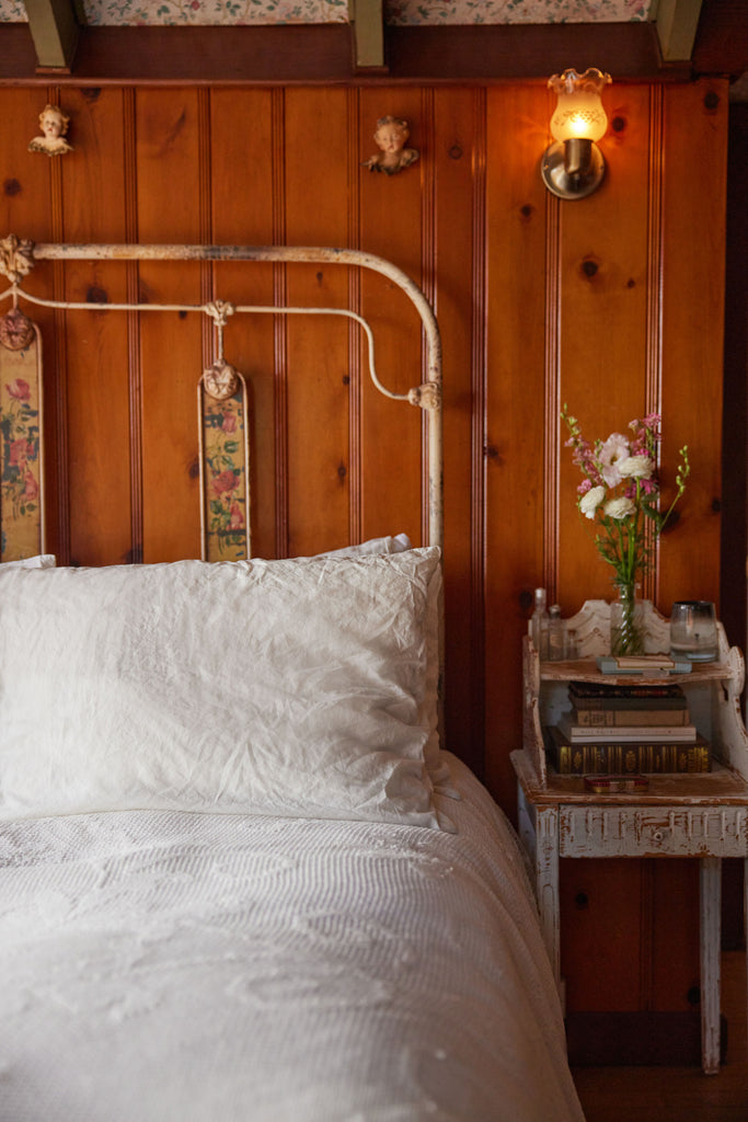 Chase Cohl's bedroom has an antique iron bed with floral inset panels and white bedding. Next to the bed is a distressed antique night stand with books and a glass vase full of flowers.