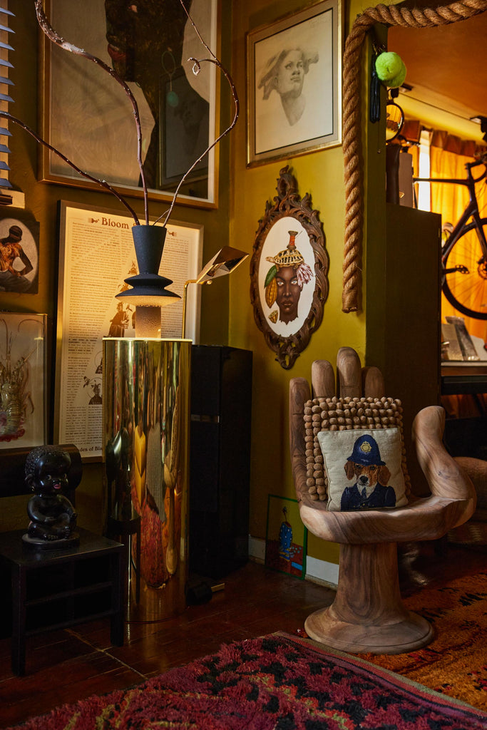 A wooden hand chair holds a textured throw pillow and pillow with a dog in a police uniform. Next to the hand is a gold illuminated tall pedestal with a uniquely shaped tall vase holding branches. Behind the chair hangs a gallery wall.