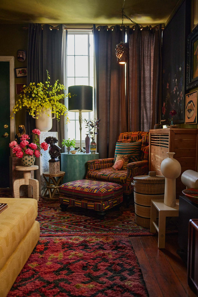 A multicolored upholstered arm chair and ottoman sits in the corner of Maurice Harris's living room. Small tables hold plants and flower arrangements to the chair's left, while a wooden dresser and woven basket sit to the chair's right.