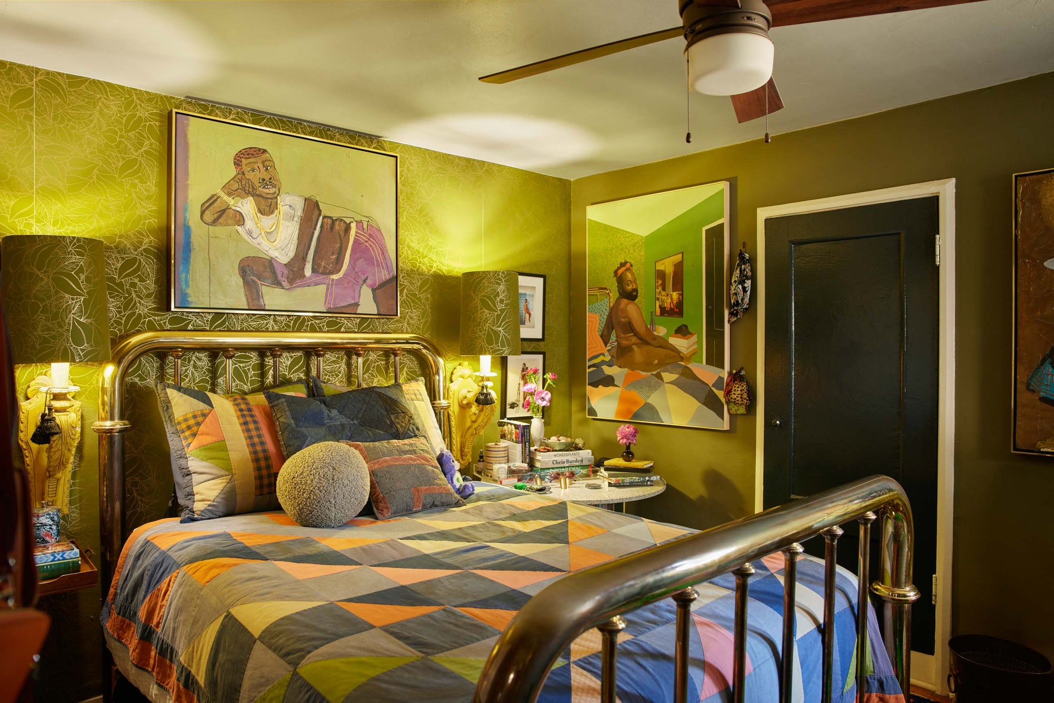 Florist and artist Maurice Harris's bedroom has a gold metal bed with a colorful quilt and pillow shams. Above the bed hangs a modern portrait of a man in a white shirt and purple shorts and to the right hangs a portrait of Maurice in his bedroom.